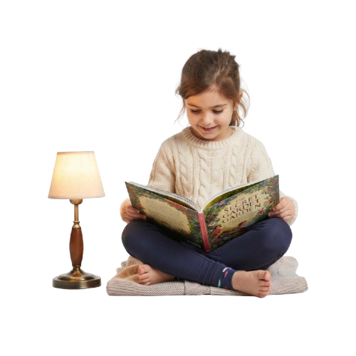 A young girl sitting on a bed reading an open book with a small lamp beside her.